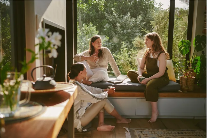 Three women sharing a conversation beside a large sunlit window.